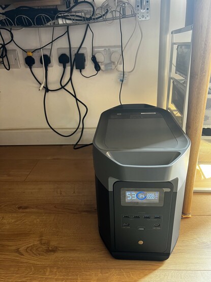 A huge, grey monster of a battery sitting on my office floor. The LCD is on showing charge status. The front is brimming with USB ports. Four type A, two type C.