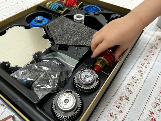 The Spintronics box open on a table. A small hand is removing one of the components- a red and gold gear switch. There are a half dozen or so other gear-style components in view and some haxagons are the bestagons metal baseplates.