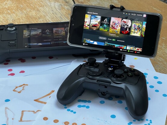 Picture of a POCO X4 phone clamped to a PowerA Moga XP-Ultra controller and phone clamp. It’s displaying the Steam Deck UI. A Steam Deck can be seen in the background displaying the same picture.