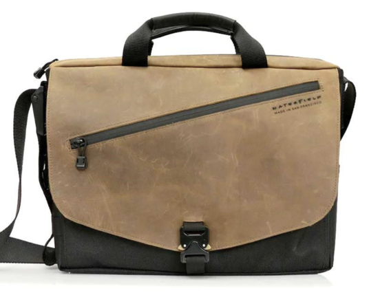 A black messenger bag with a brown leather flap. It has a metal buckle and a jaunty diagonal zip pocket.