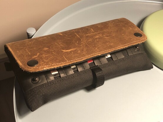 A black mesh case with a weathered brown leather flap. A few PS Vita games and game slot to SD card adapters are visible in little pockets peeking out under the flap.