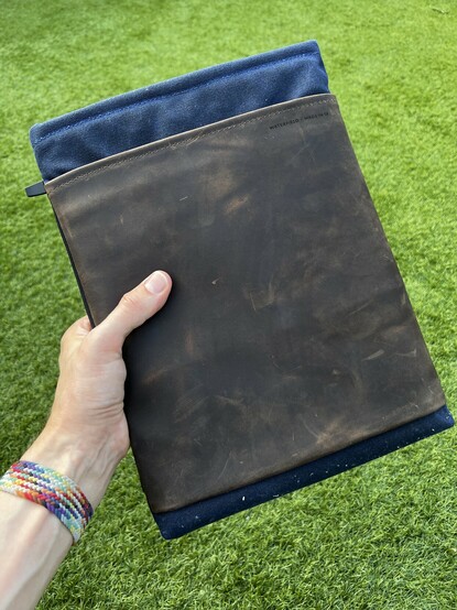 A blue waxed canvas laptop sleeve accented by a large, weathered, chocolate leather front pouch. I’m holding it up outside in front of a green lawn.