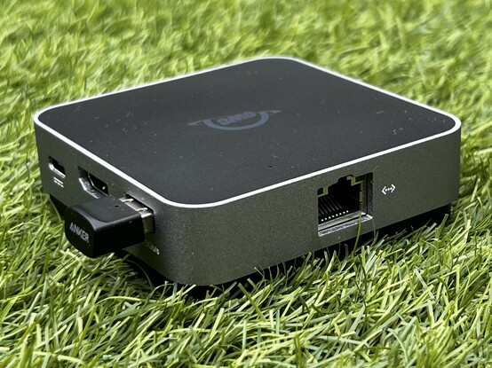 The OWC Travel Dock E sat on some grass. It’s turned 45 degrees to the camera, showing two edges. One edge has a USB Type-C, HDMI and USB port and the other an Ethernet port.