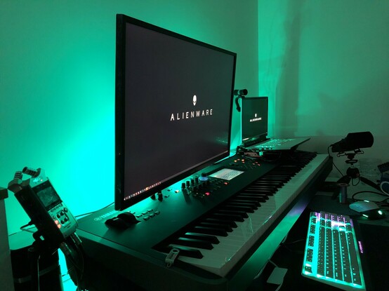 A desktop lit up greeny teal by some hidden lights. There’s a huge keyboard/piano occupying most of the scene. A lit up computer keyboard is sat in front. There’s a microphone in the background and a field recorder in the foreground. It looks like I’m set up to do serious musical business.