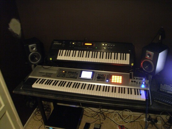 A dark blurry photo showing a large metal piano lit up with a smaller black synth mounted above it. There are some studio monitors. There’s a glow of an unseen monitor in the right edge.