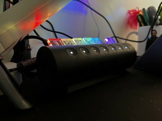 A rounded USB hub lit up with rainbow colours. It’s on a desk with a monitor arm above it and a mug in the background with what looks suspiciously like my face on it.