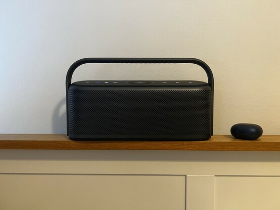 A radio-like Bluetooth speaker sat on the headboard of a bed. It’s grey, minimalist and curvy. The handle shows just a hint of hand grip and there are illuminated buttons just visible on top.
