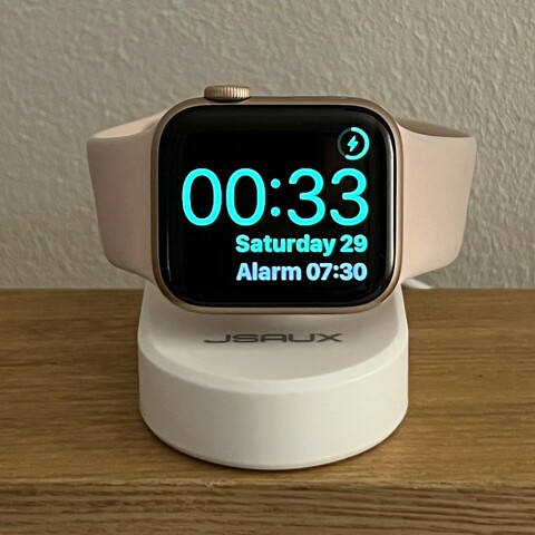 An Apple Watch with a pink sports band magnetically clasped onto a little white charger. It’s tilted upright so you can see the time and date easily.