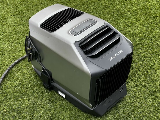 The EcoFlow WAVE 2 sat outside on grass. It’s a chunky grey and black plastic air conditioner with two vents on top and a little LCD display with controls. The battery sticks out underneath like a little chin, giving the impression that it’s a robot head.