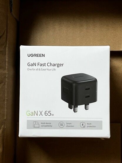 A closeup of a cardboard shipping box. Inside is the white box of the GaN fast charger. It has some holographic text reading “GaN X 65w”. The plug - pictured - is plain black and shows two stacked USB C ports.