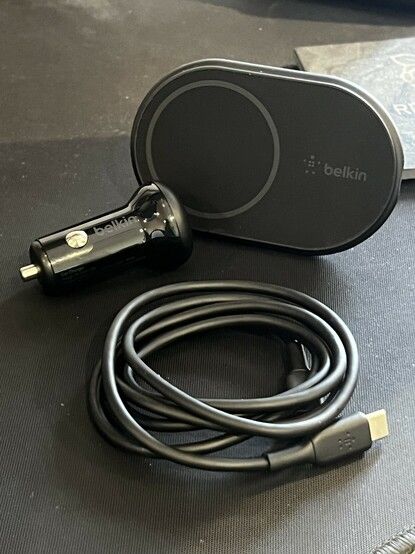 Photo of the Belkin car charger, a matte, charcoal grey oval shaped pad. Along side it is the USB cable and a gloss black 12v car power supply. The “lighter socket” type.