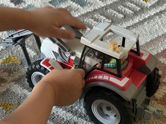 Small hands plugging a little green toy electric charger cable into the side of the tractor. It’s clear how little of the toy the vibrant red covers here, with most of it grey and white.