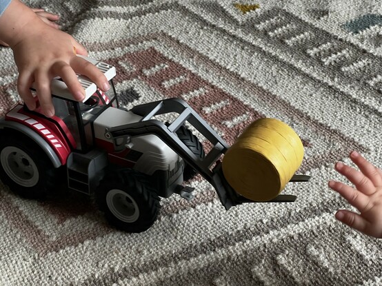 A photo of Playmobil’s electric tractor being pushed along the floor by a child’s hand. Another, smaller hand reaches into the right of the shot; trying to grab the tractor. It’s coloured… you guessed it … red, white and grey.