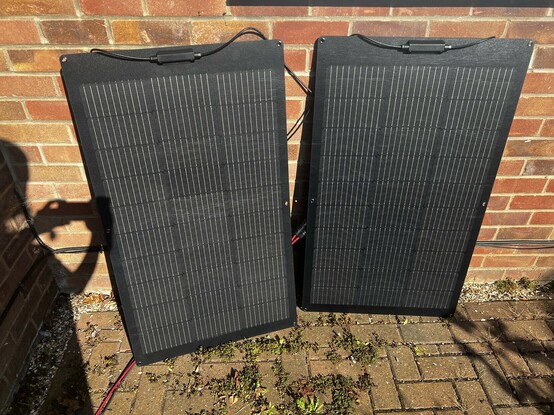Two EcoFlow 100W flexible solar panels leaned awkwardly against a wall, standing on their short edge. They stand up surprisingly well. On the left you can see a harsh shadow of me taking the photo. Red and black wires trail across the ground.