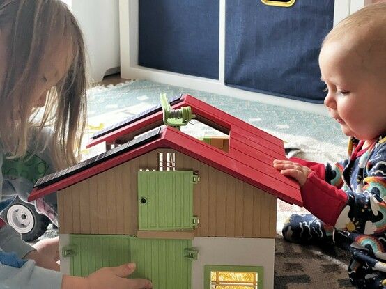 A baby and a toddler playing with the Playmobil large organic farm. It has a vibrant red roof with two rows of solar panels. The roof is cut-away in the middle to allow access. There are muted green doors visible at the end. There’s a small green gantry crane peeking out of the top. The look of concentration on babies face is the precursor to him tipping the whole farmhouse over.