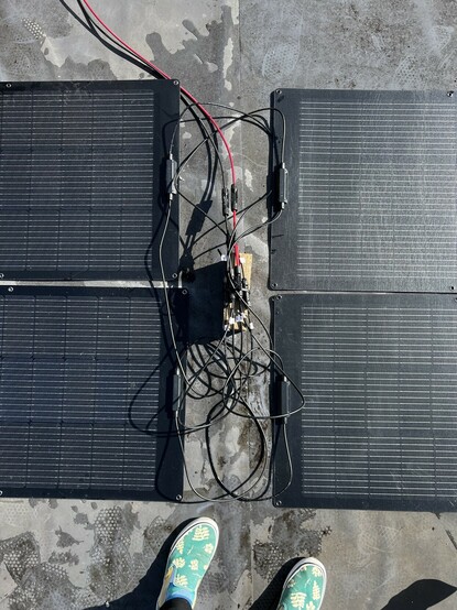 Four solar panels arranged in a 2x2 grid with a nest of cables between each pair. The cables coalesce on a regular house brick to which they are secured, a black and red cable snake out of the top of the image. My green shoes can be seen poking into the bottom.