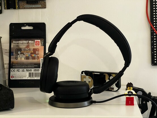 A pair of on-ear headphones stood upright with one of the ear cups in a wireless charging pad. A little Lego figure is holding up the charging wire. It’s supposed to be me, but the hair is wrong.