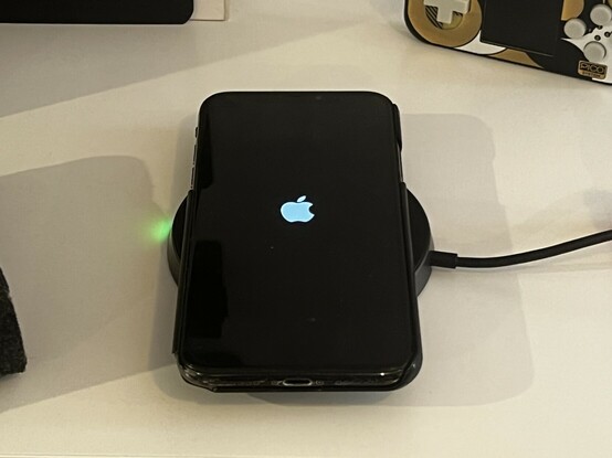 An iPhone X in a very shabby plastic case sat on top of the wireless charger. It’s just turned on and showing an Apple logo. A little green LED is illuminated on the charger.