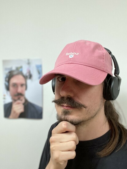 A headshot of me - a moustachioed man - wearing a pink baseball cap with the Evolve2 Flex headphones over the top. I’m trying to strike a thoughtful, beard stroking pose. In the background is a printout of a photo of me wearing the headphones with no cap and striking the same pose.