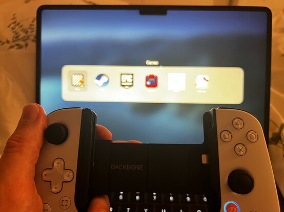 My hand holding a black and white controller in front of a blurred Mac laptop screen. There’s a conspicuous recess where a phone would otherwise be.