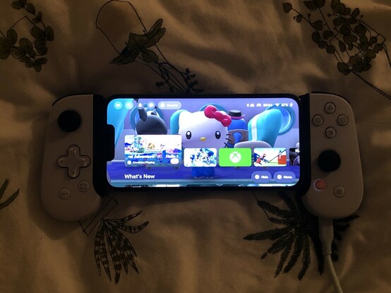 My iPhone in a white game controller grip sat atop my bed over. The phone is showing the Backbone App Home Screen with Hello Kitty selected.