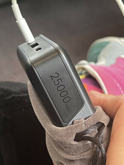 A side view of UGREEN’s portable battery showing “25000mAh” text on the side. The design is straight forward silver with the ports, LCD and labelling occupying a wrap-around black area that cuts into the top of the device. There are two USB Type-C ports and one Type-A. My pink and purple socks and pink, baby blue and green shoes are visible blurred out in the background.