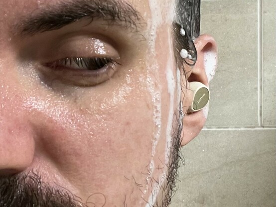 A close up photo of the left side of my face and left ear. Shampoo suds are running down my cheek and my hair is wet. Poking out of my ear is a small beige earphone with a flat top. It has “Jabra” written on it.