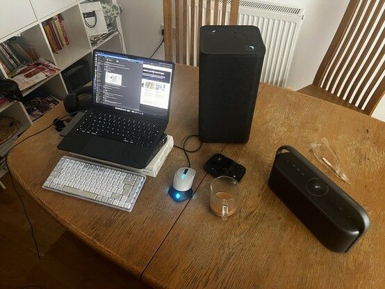 Looking down on a worn old oval dining table. On the table is a large, black, obelisk of a Bluetooth speaker and another sleeker speaker in a more radio style. There’s a glass mug with the dregs of tea. A Razer Kishi folding phone gamepad, folded up. A white and black mouse with blue lights. A computer mouse… that is. On the left is a laptop stood atop a couple of boxes. In front of it a white and grey wireless keyboard.