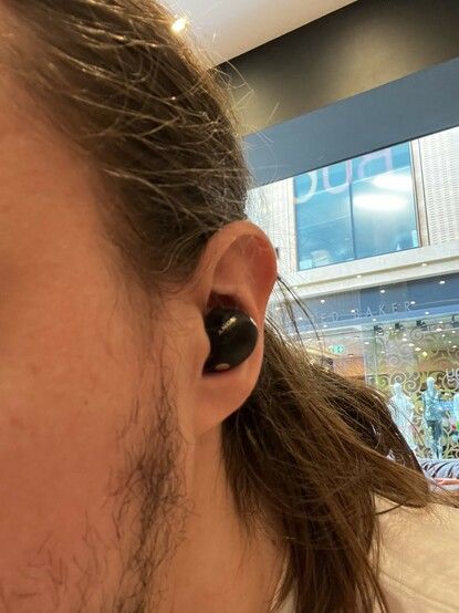 A slightly blurred picture of my left ear with the little black and gold earbud picking out. It’s quite large, filling the inner part of my ear. My long hair- only somewhat tamed by a tie- trails back over my shoulder. In the background is the front of the Cambridge Ted Baker store.