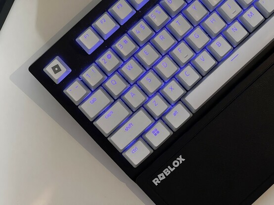The left-hand edge of the Roblox branded Razer keyboard, placed at 45 degrees showing approximately half the main keyboard. There’s a Roblox text logo on the wrist-rest and a Roblox icon logo on the Esc key in the very corner.