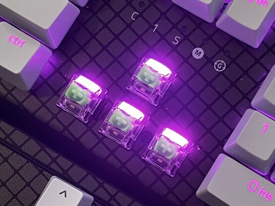 A more top down view of the arrow keys. The text “C 1 S M G” is sulked above, denoting the function of the keyboard status LEDs. The key caps show big, bright LEDs either the full width of the cap or well diffused. They are lit up purple, but are so bright it looks white toward the centers.