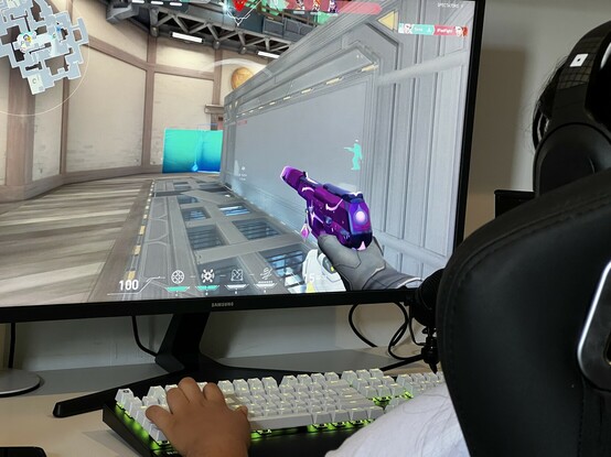 An over-the-shoulder shot of a large screen showing a cartoon-styled videogame- probably World of Warcraft or Candy Crush. In the foreground the corner of a Noblechairs chair can be sent, and over the top a head wearing the Roblox branded Razer headphones. A hand reaches out to the QWOP keys on a black gaming keyboard with white keys, lit up a sickly chartreuse.