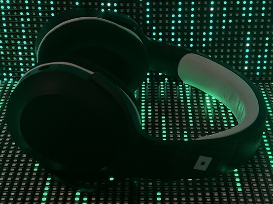 A pair of black and white headphones sat on top of a pair of RGB matrix panels and in front of another. They are lit up with green glare. The Roblox logo is picked out in white on the side of the headband. The padded inner part of the headband is white. There’s white piping between the ear cups and pads.