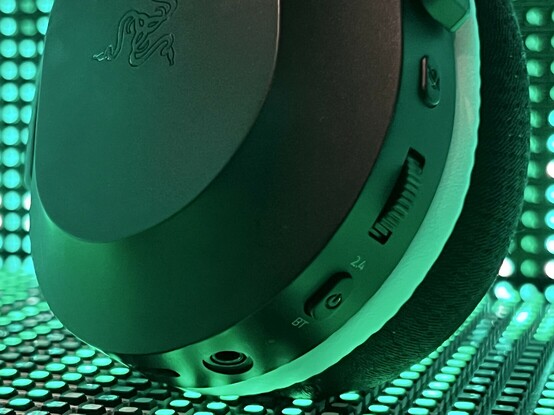 A close up of the left ear cup of the Razer Barracuda X. It shows the white that splits the main ear cup from the padding, plus- from left to right- the USB type C port, 3.5mm jack, 2.4GHz/Bluetooth and power button, volume wheel and microphone mute. The second 3.5mm socket for the microphone is the opposite side of the cup.