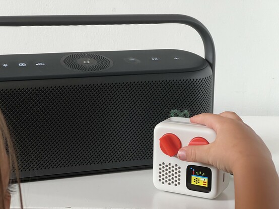 A radio-like Bluetooth speaker sat on a white table. In front and to the right is a Yoto Mini, a small, white, cuboid kids audio player with two orange button dials, a speaker grille and a tiny display on its front. A small hand is reaching over to press one of the dials. A fun pixel art picture of a radio is shown on the display. There’s also a blue dot in the very top left, indicating that a Bluetooth device is connected.