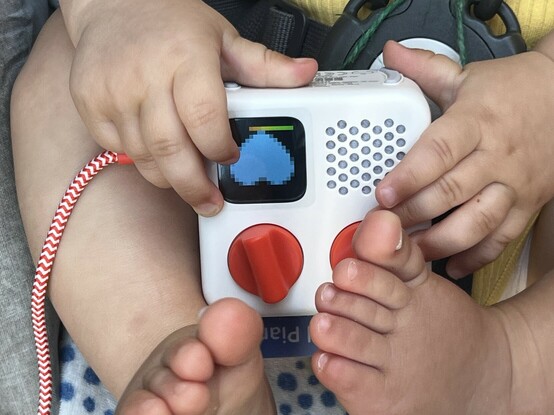 The hands and feet of a baby, clutched around the Yoto mini audio player. It’s upside-down with an orange and white braided USB cable poking out of the left. There are two vibrant orange knobs on the front, a small speaker grille and a screen displaying a heart.
