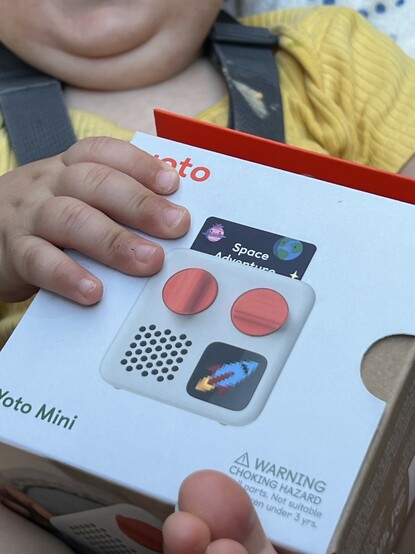A tiny box being held by a tiny baby hand. It’s orange and white, the name “Yoto” is partially obscured by a finger. On the front of the box is a photo of a small, square audio player with a small screen, two orange dials and a speaker grille.