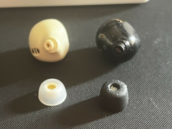 A blurry closeup of the Jabra Elite 7 Pro (left) and the Sony XM5 (right) earbuds. They are each sat with their respective ear tip, detached and placed in front. The silicone tip of the Jabra earbud is pristine, while the foam tip of the Sony is grungy, worn and grubby.