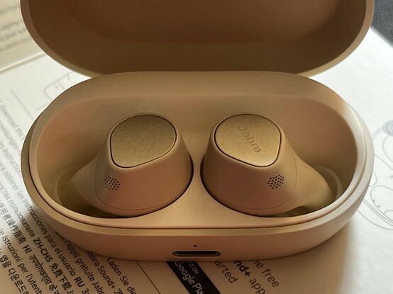 A photo of the Jabra Elite 7 Pro earbuds sat in their - open - charging case. The lighting is warm, giving them a warmer look than their beige and gold exhibits in the flesh. There’s a USB C port in the front of the case. The front!
