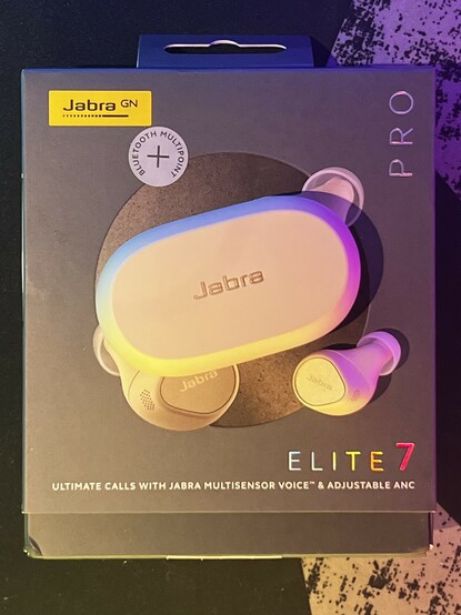 The Jabra Elite 7 Pro charging case and an earbud lying atop the box. They are lit boldly with rainbow light from offscreen, giving them a colourful, pastel look. The image lacks depth so - without noting the harsh shadows on the items atop the box - it might be confused as merely a picture of the box art.