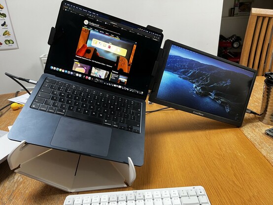 A photo depicting a 14” auxiliary screen clamped to the back of a laptop. It’s weight has tipped the laptop over on its desktop stand, causing the whole thing to sit at a jaunty angle with the corner of the auxiliary screen touching the table top.