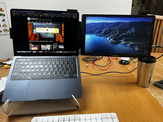A laptop with an auxiliary screen clamped onto the back. Since the laptop is raised up off the desk with a riser, there’s a coffee cup placed under the corner of the auxiliary screen to hold the extra weight and stop it tipping over.