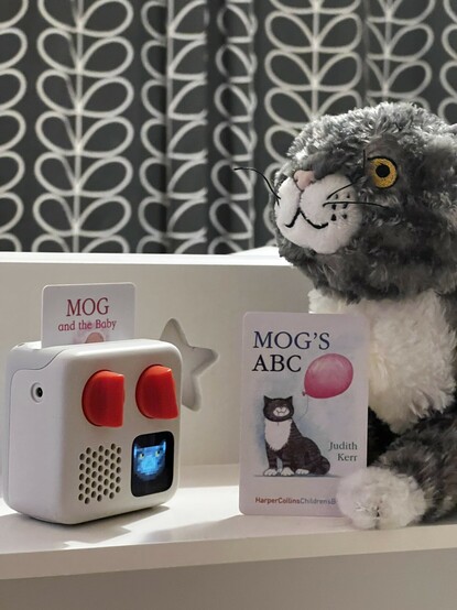 A shot of a shelf, with a grey and white cat plushie poking in front the right-hand side and propping up a credit card sized card with “Mog’s ABC” written on it and a picture of the very same cat. On the left is a tiny white, grey and orange audio player with a similar card inserted entitled “Mog and the baby.” On its screen is a little pixel art facsimile of Mog, the titular cat.