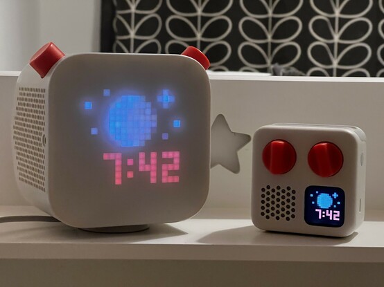 A photo of two similarly coloured audio players, both grey with orange accented dials, both showing a moon and star pixel art on their respective displays, along with the time: 7:42. The larger of the two sits on a charging base and a wire trails off out of shot on the left. Behind them is a star-shaped cut-out in the wooden shelf, and in the background are slightly blurred out dark grey curtains with a leafy stem pattern.