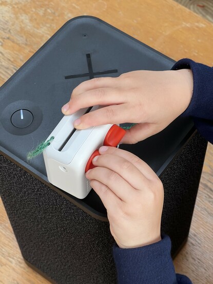 A photo of small child’s hands operating the orange dial on a mini audio player. It’s sat atop a huge, Bluetooth speaker.
