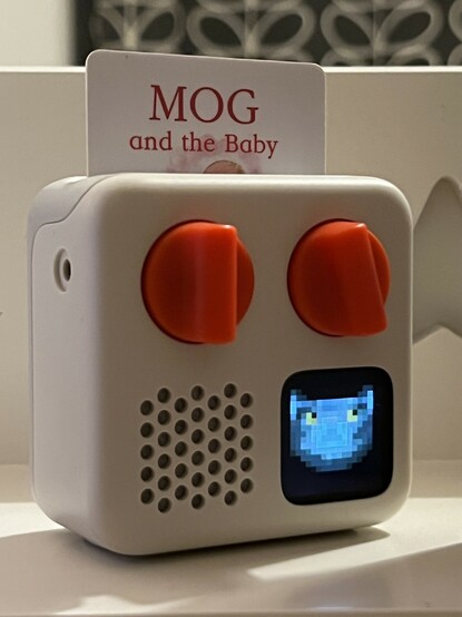 A tiny kids audio player with two bright orange dials on the front occupying the top half of the device. In the bottom right corner is a display showing pixel art of a grey and white cats face. In the bottom left corner is a speaker grille. On the side is a hole- a 3.5mm audio jack- and out of the top peeks a credit-card sized card entitled “Mog and the baby.”