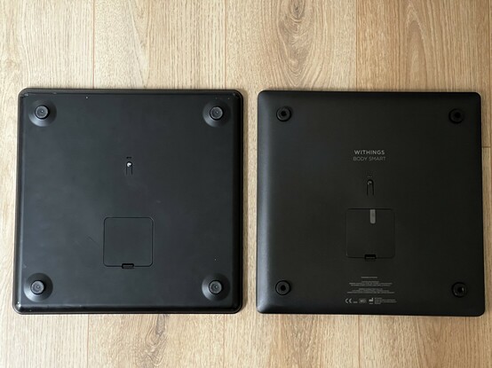 Two sets of bathroom scales turned upside down and laid side by side. They are very similar, with the same style battery compartment and pairing button. The newer one on the right has redesigned feet that have a small locating hole in the middle for carpet load spreaders.