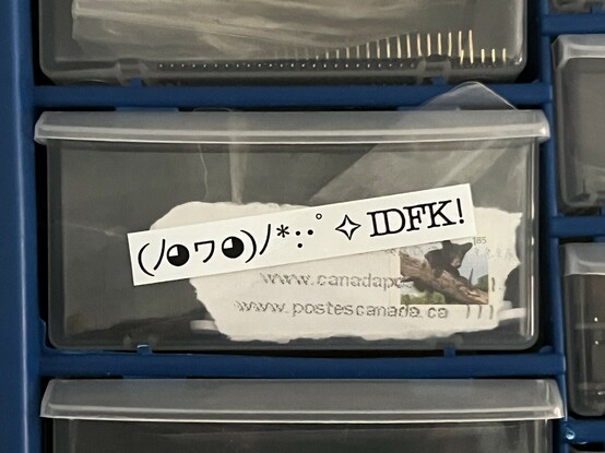 A component drawer with a shred of envelope bearing a Canadian stamp pushed to against its front by whatever junk lies therein. A label on the front has a Unicode rendition of a little cute face throwing stars and sparkles ending in the letters “IDFK” - or “I don’t forking know”