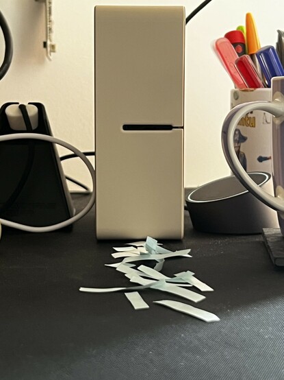 A photo square on the front of the label maker. It’s stood on a desk with a mug of pens in the background. At its base is a smattering of paper offcuts and backing paper from labels.
