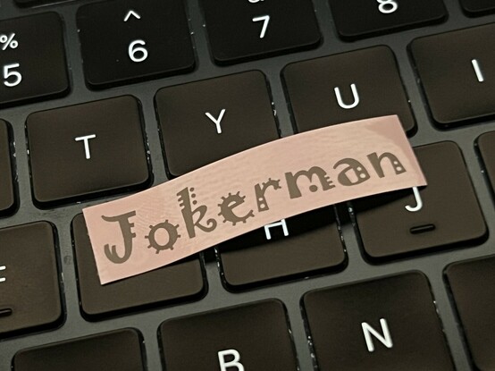 A small label resting upon a laptop keyboard. It reads “Jokerman”, and is rendered in a very busy font with lines and dots decorating its quirky letterforms.